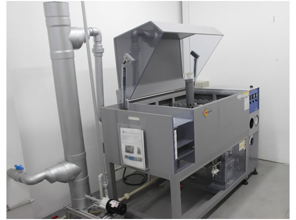 Cámara industrial de pruebas de corrosión y niebla salina con tapa basculante transparente abierta, mostrando la torre de atomización en el interior del tanque de prueba. El equipo cuenta con un panel de control a la derecha y un sistema vertical de tuberías de ventilación gris a la izquierda.